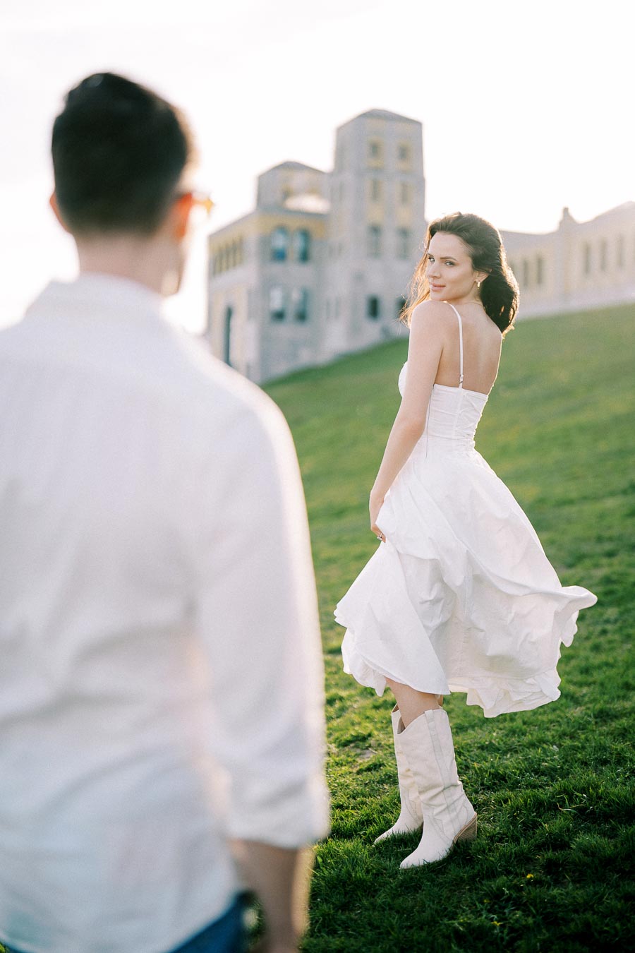 A woman in a white dress and boots smiles and looks back at a man on a grassy hill, with a historic building in the background under bright sunlight.