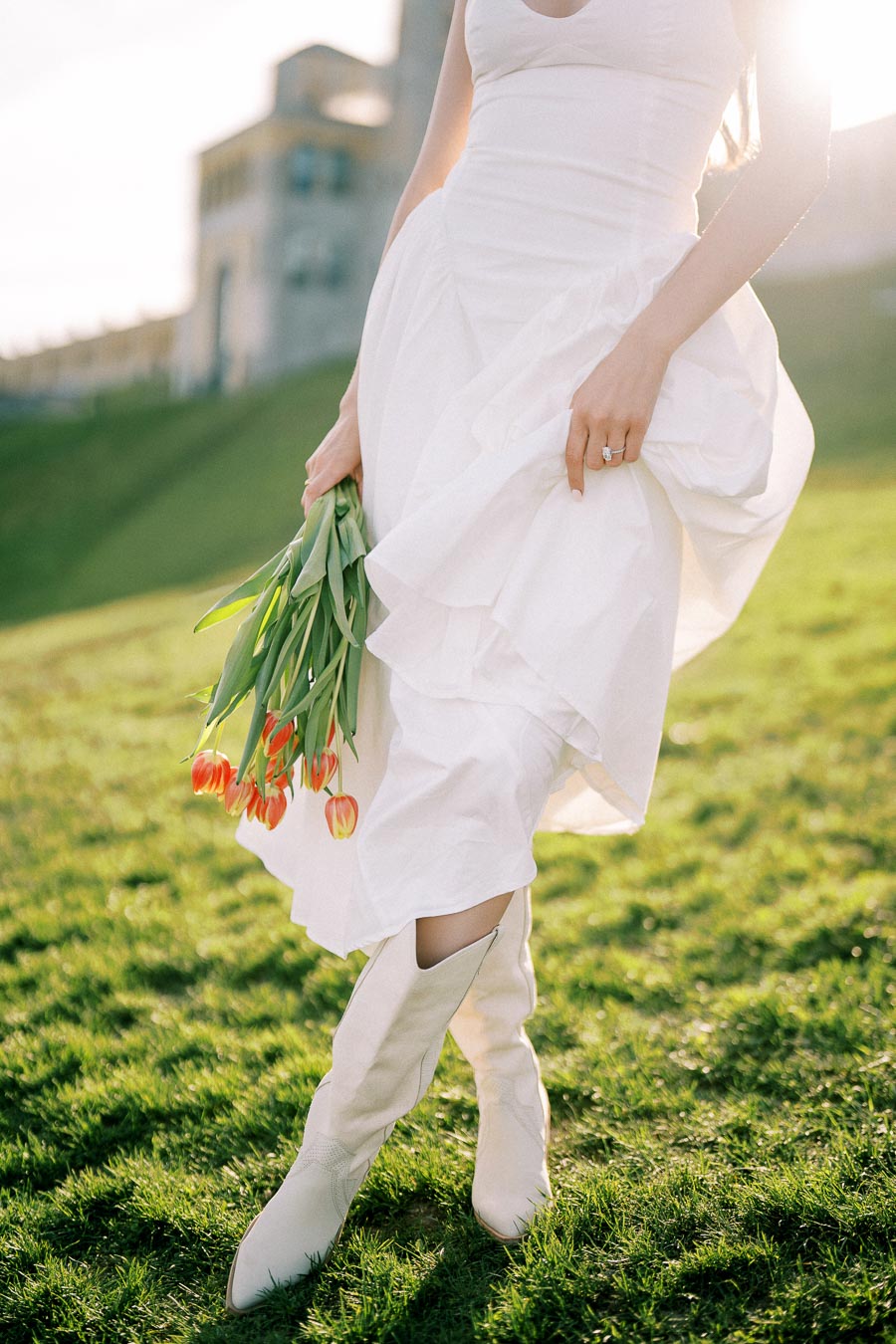 A woman in a flowing white dress and white boots holds a bouquet of orange tulips while walking on a sunlit grassy hill, with a blurred historical building in the background.