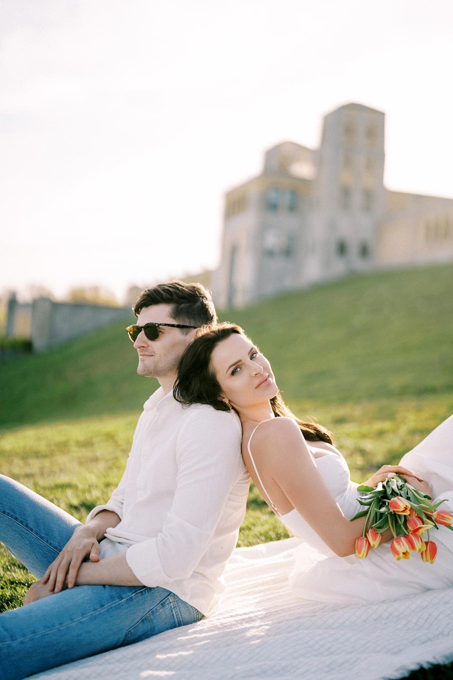 A couple relaxing on a picnic blanket in a sunny park, with the woman holding a bouquet of tulips and a historic building in the background.