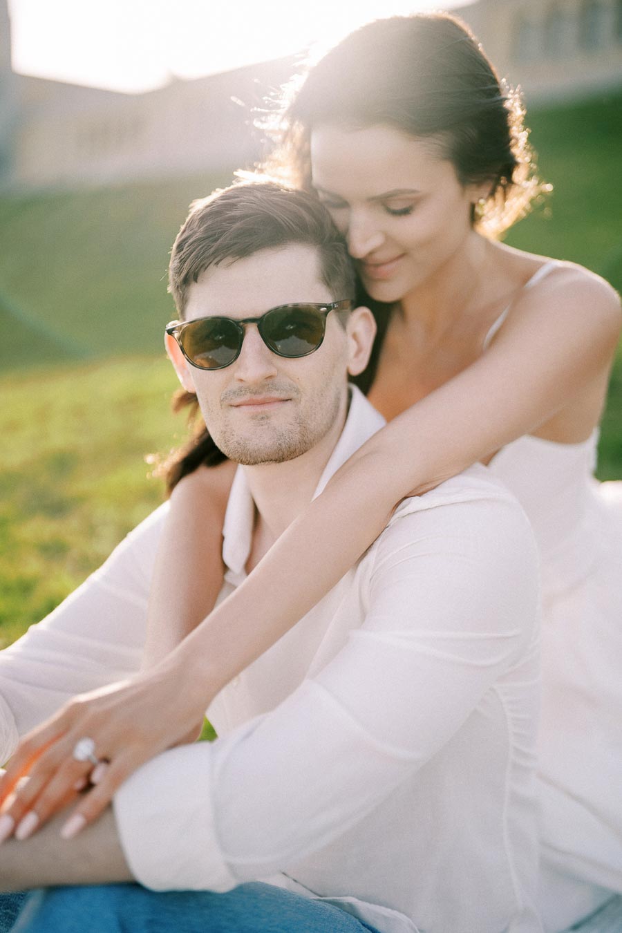 Romantic couple embracing outdoors, woman in white dress hugging man wearing sunglasses, sitting on grassy hill with sunlight in background.