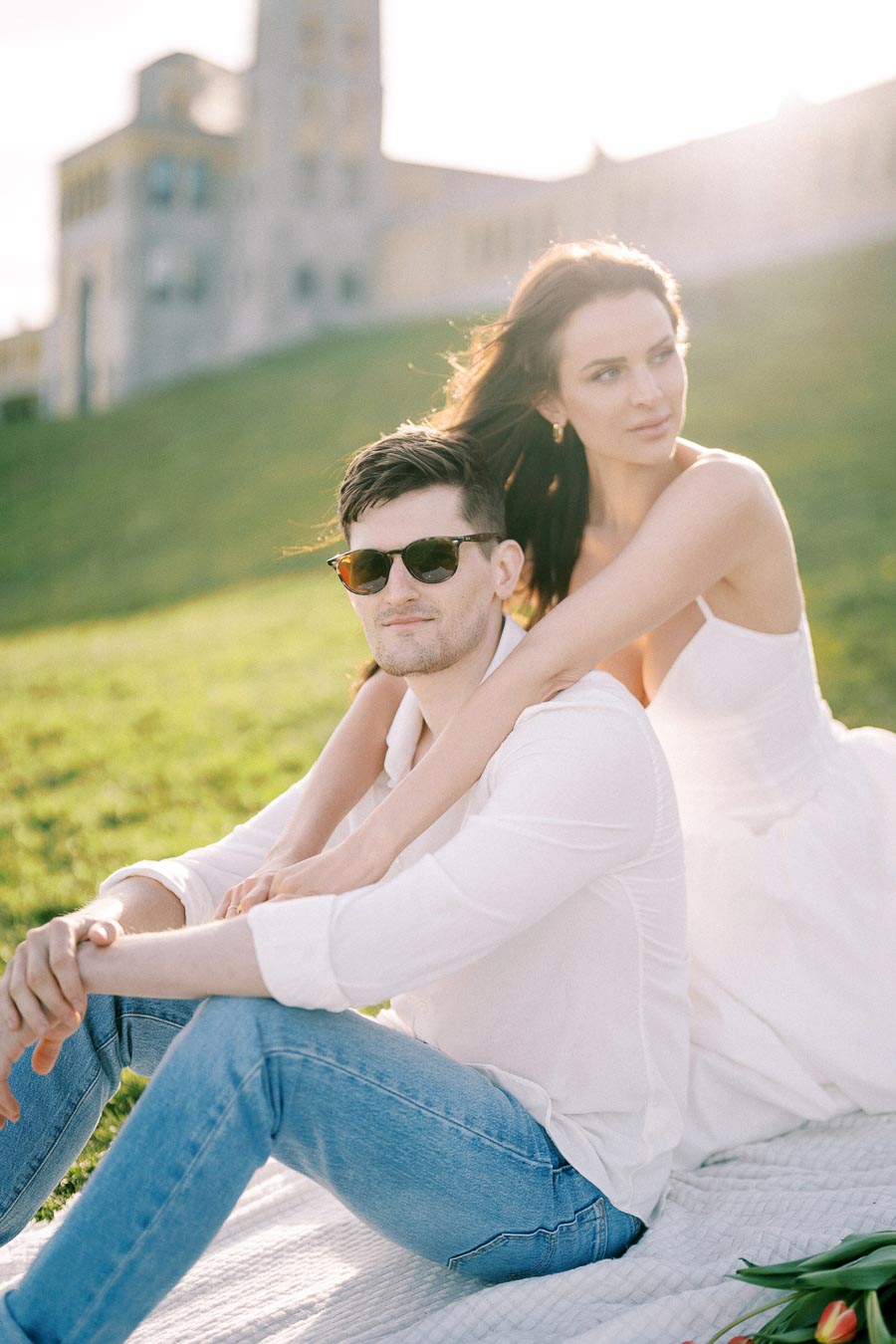 A couple enjoying a sunny day in the park, with a woman embracing a man from behind. The man wears sunglasses and a white shirt, while the woman is dressed in a white dress. They are sitting on a blanket with a grassy lawn and a large building in the background.
