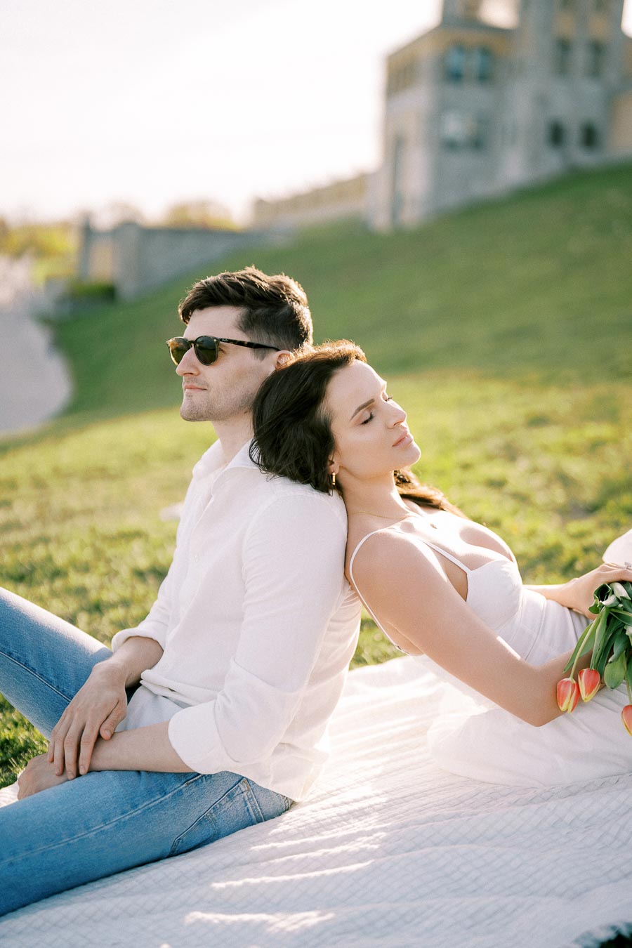 A couple enjoying a sunny day at the park, leaning against each other on a blanket. The woman is holding tulips, and both appear relaxed and content.
