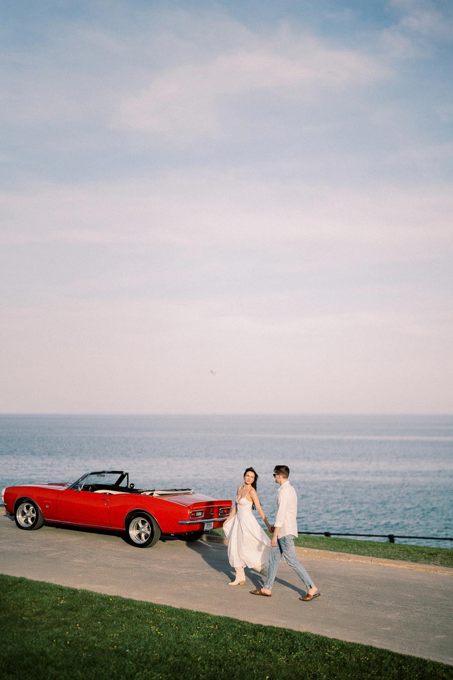 A couple walking hand in hand along a coastal road next to a red vintage convertible car, with a serene ocean view in the background.