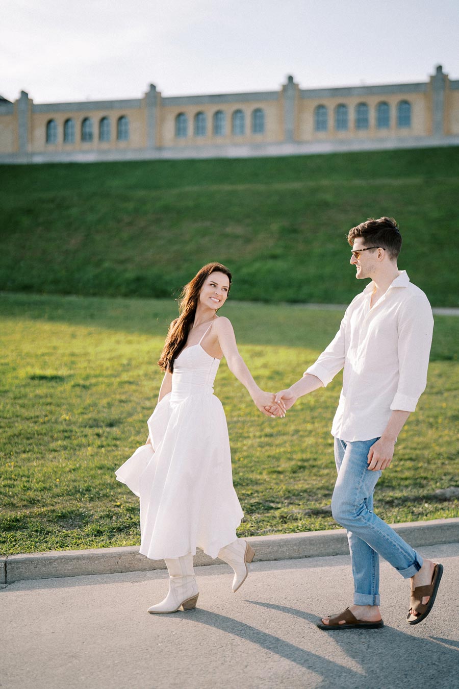 A couple holding hands and smiling while walking on a sunny pathway, with a grassy hill and building in the background.