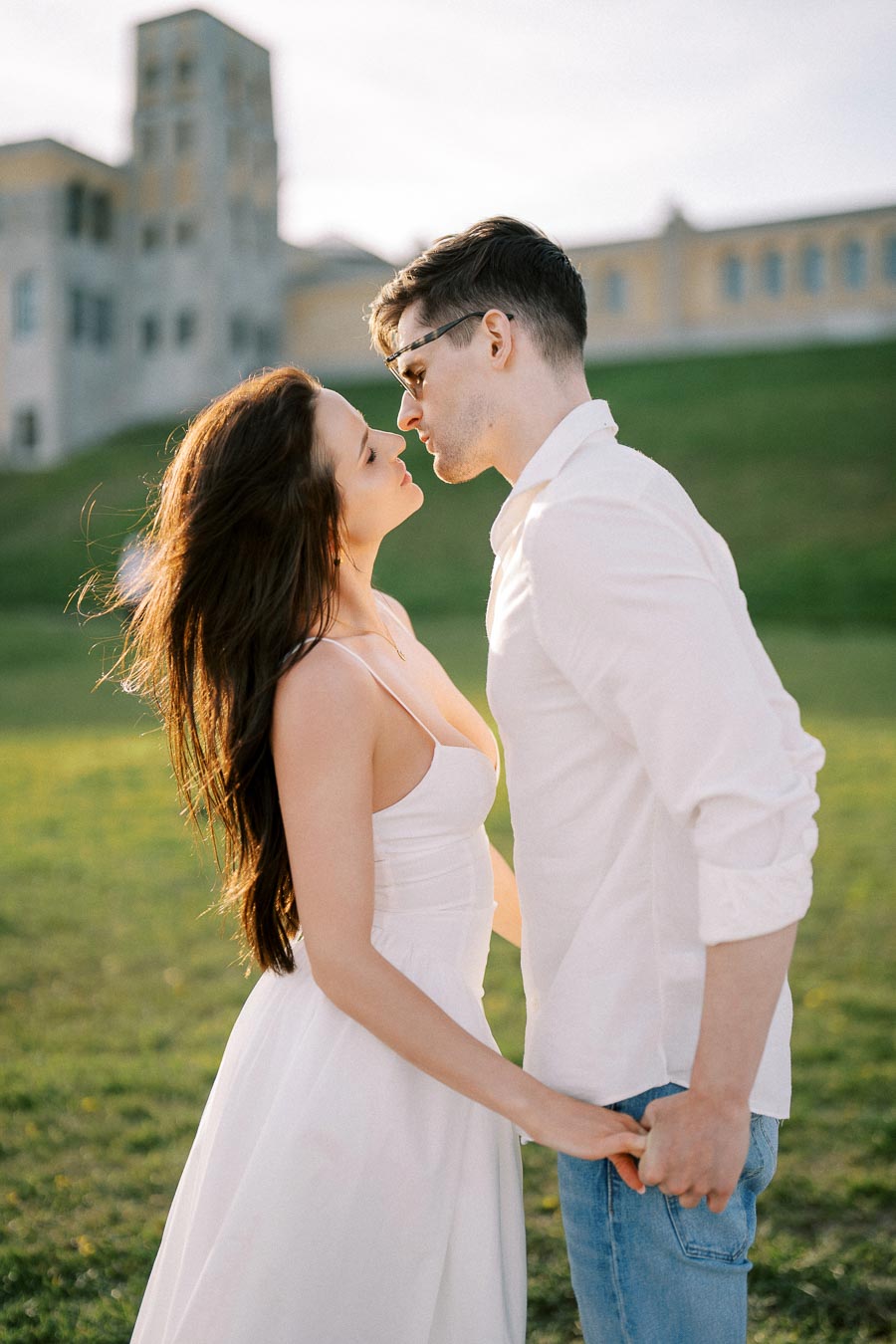 A couple in a loving embrace outdoors, with the woman in a white dress and the man in a white shirt and jeans, holding hands in a green field near a building under a clear sky.