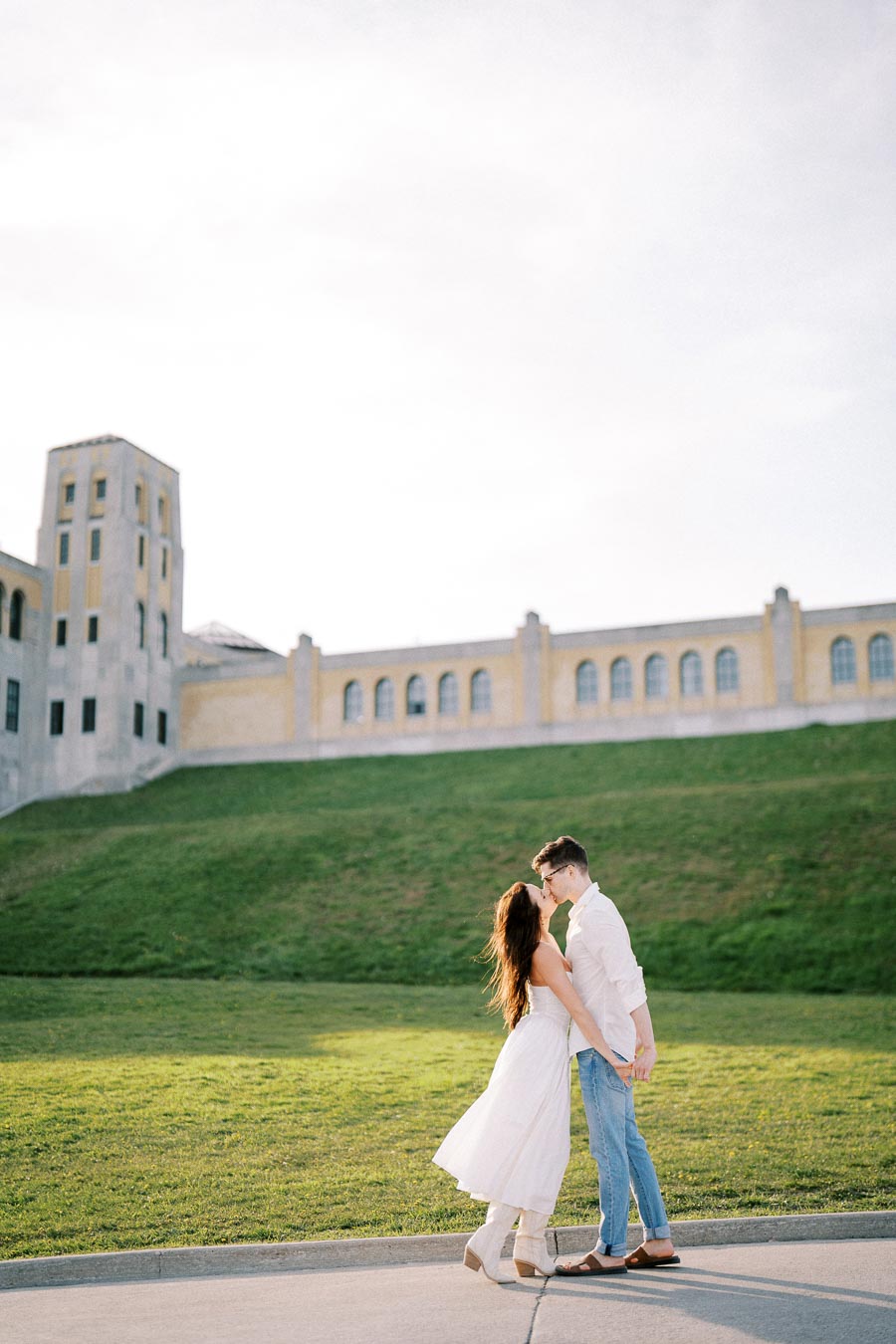 A couple embracing and kissing in front of a historic building with a large grassy area, under a clear sky.