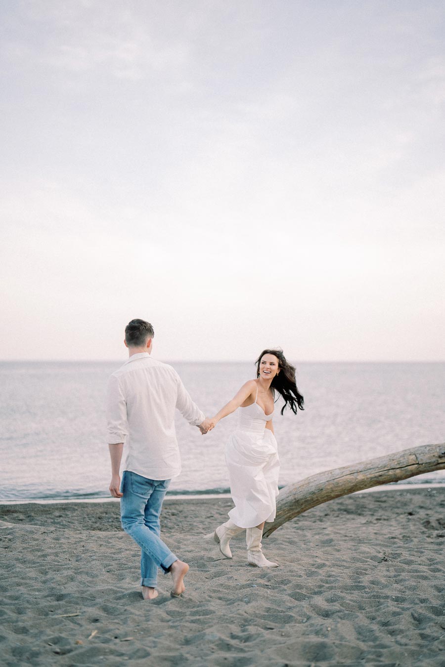 A couple holding hands and walking barefoot on a sandy beach near the ocean, with the woman smiling and wearing a white dress, creating a romantic seaside moment.