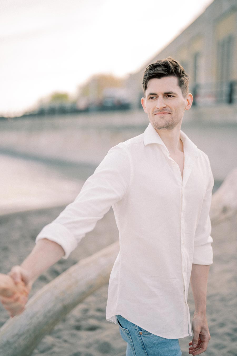 Man in a white shirt smiling and holding hands, walking on a beach with blurred background of a promenade.
