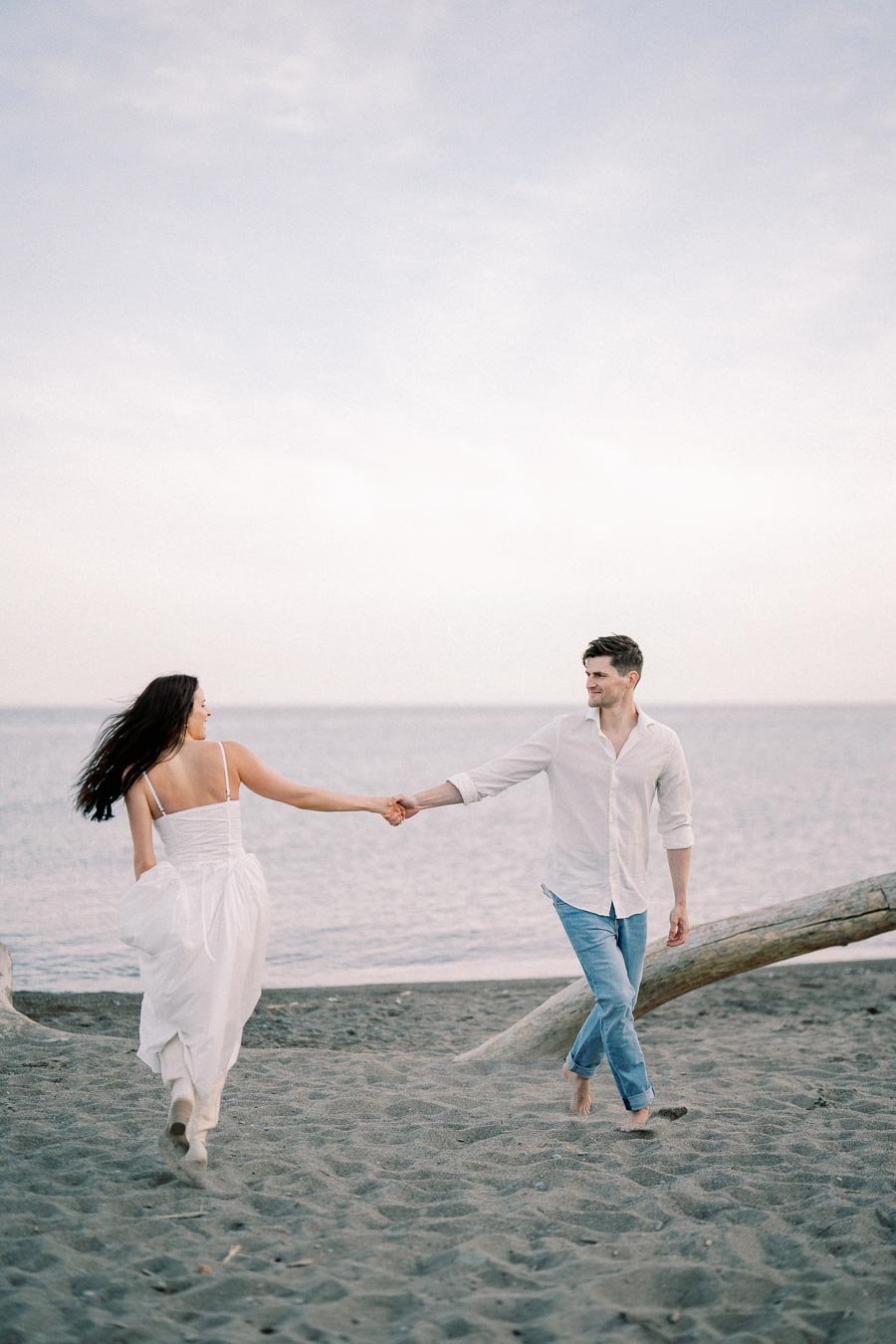 A couple holding hands and walking barefoot on a sandy beach, with the ocean and a cloudy sky in the background. The woman is wearing a white dress, and the man is wearing a white shirt and jeans.