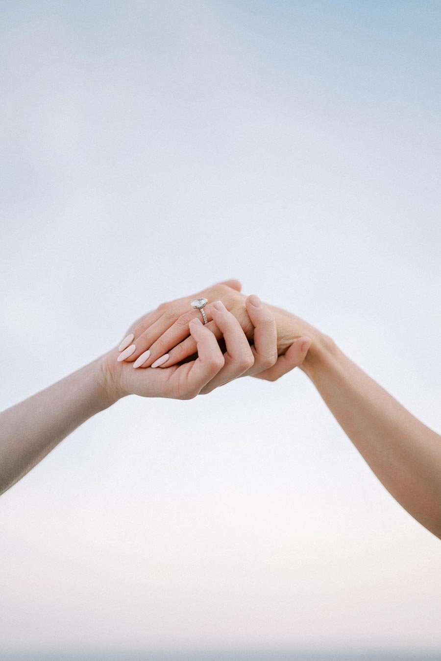 A close-up of two hands gently holding each other, with one hand wearing a diamond engagement ring, set against a clear blue sky.
