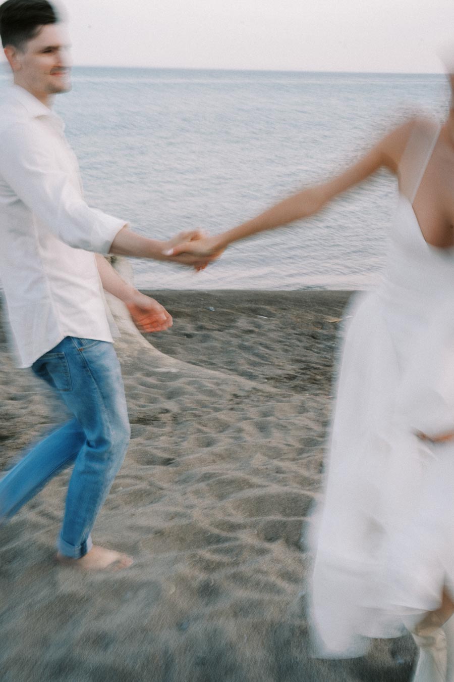 Blurry photo of a couple holding hands and playfully running on a sandy beach near the ocean, wearing casual attire and wedding dress.