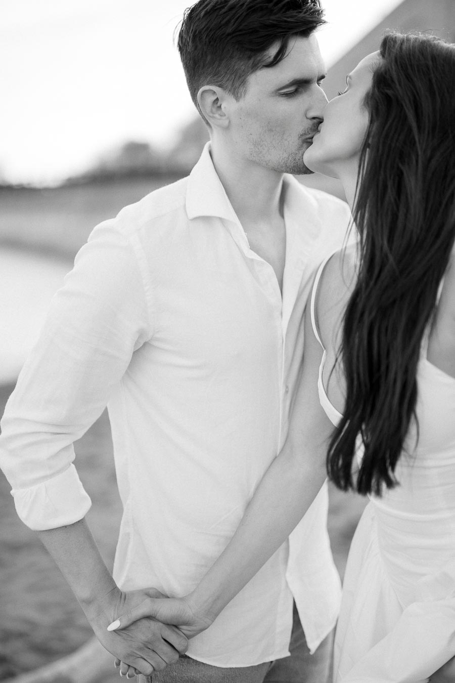 A black and white photo of a couple sharing a romantic kiss on a beach, holding hands and wearing casual white attire.