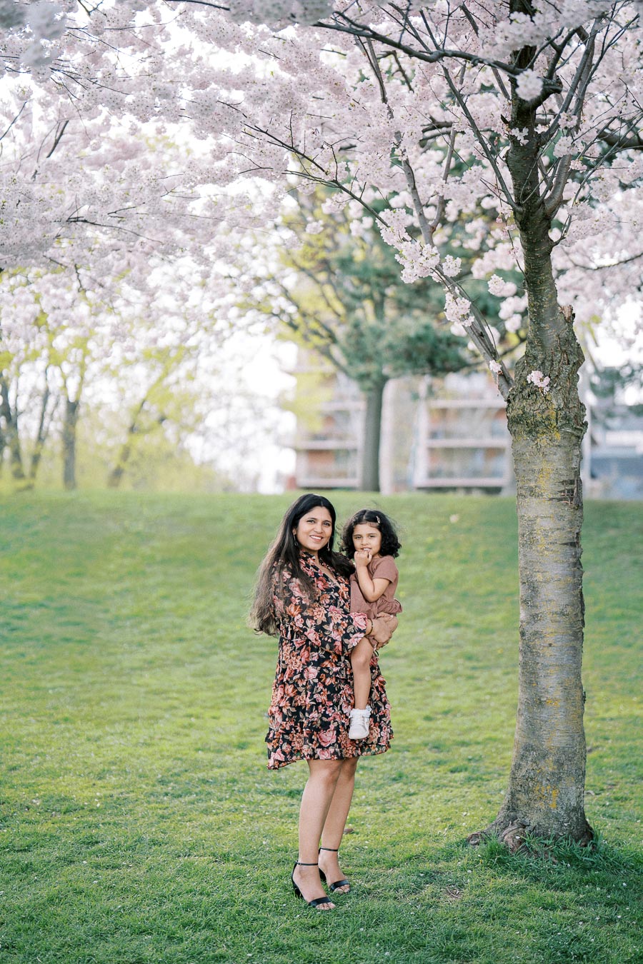 A woman holding a young child in a park with cherry blossom trees in full bloom, wearing floral dresses, enjoying a sunny spring day.