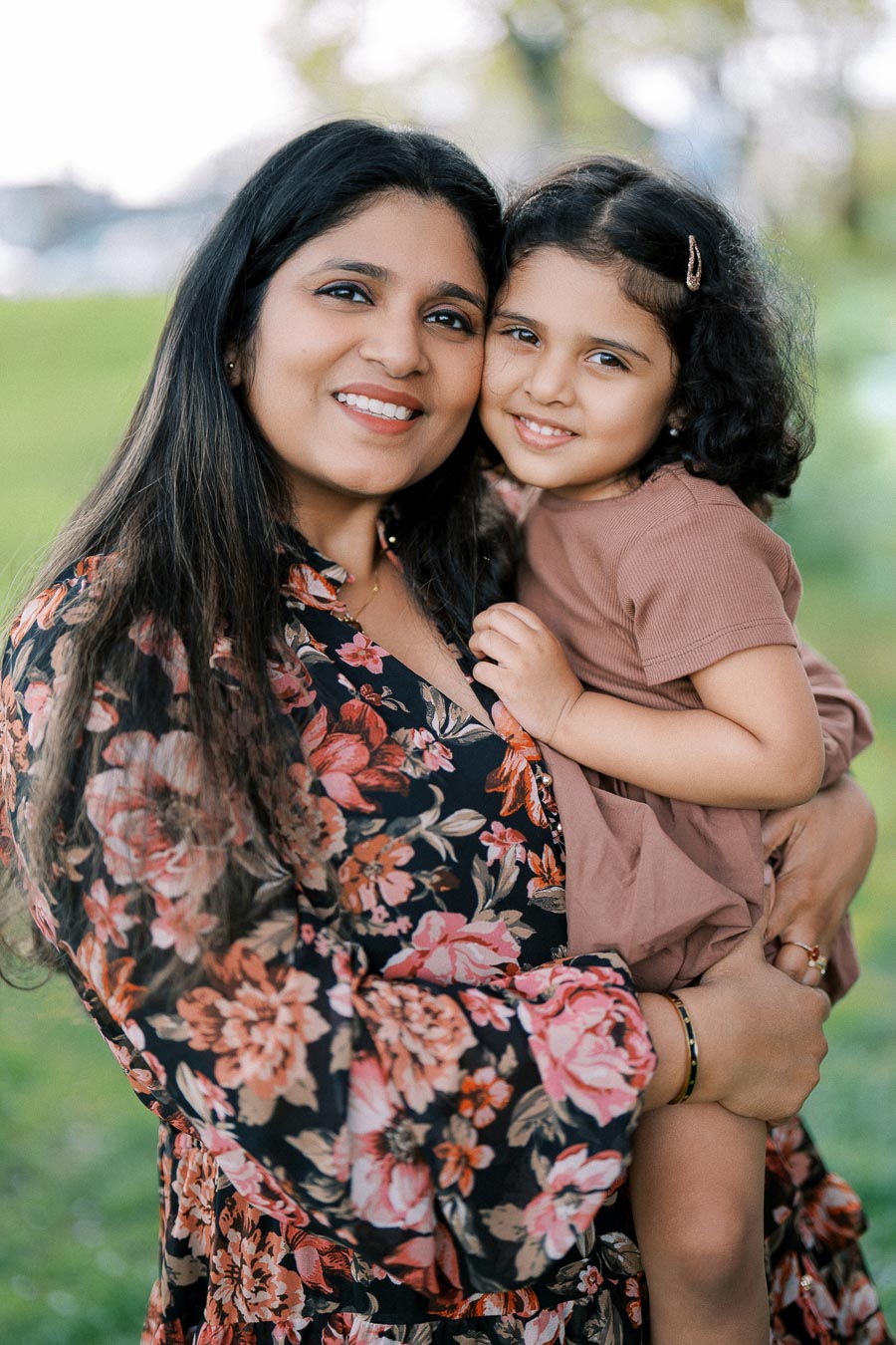 A woman in a floral dress smiling and holding a young girl in a park setting.
