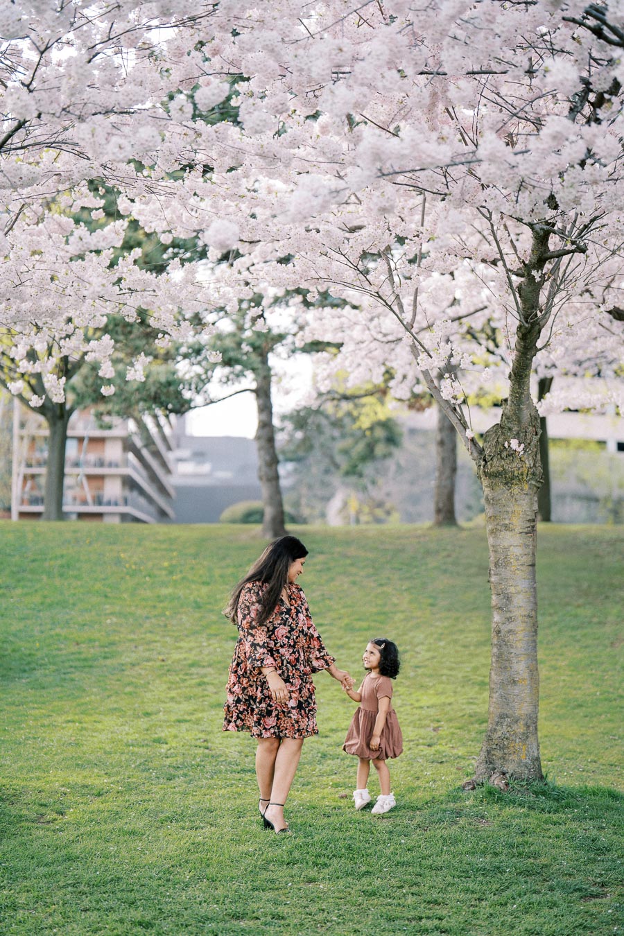A woman and a child holding hands and smiling under blooming cherry blossom trees in a sunny park, showcasing a joyful spring moment amidst vibrant greenery.