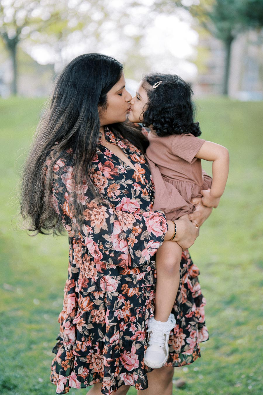 A mother in a floral dress lovingly holds and kisses her young daughter in a park setting, surrounded by lush greenery.