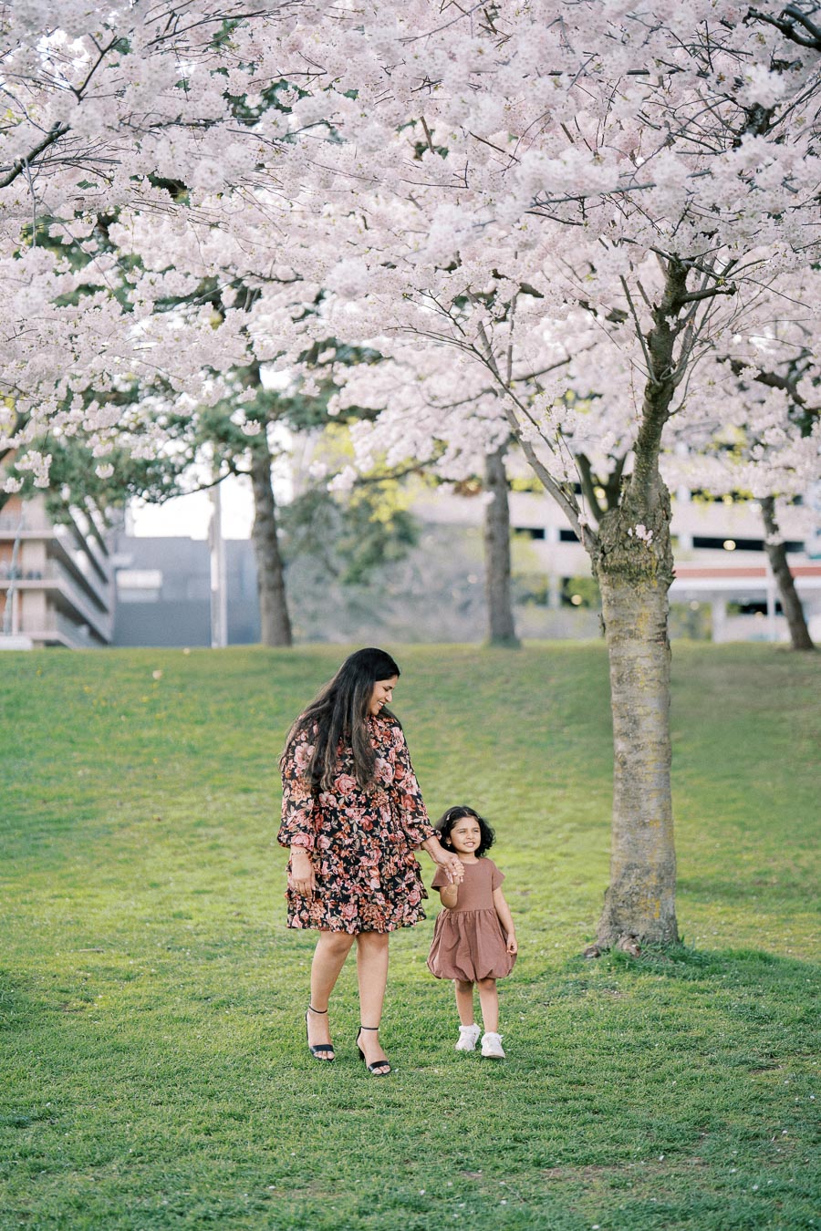 A woman and child walking hand in hand under blooming cherry blossom trees in a park, dressed in matching floral outfits.