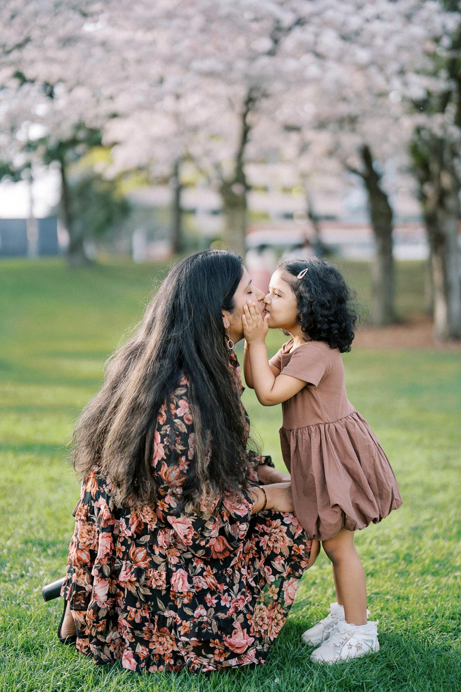 A mother and daughter share a tender moment under blossoming cherry trees in a park. The mother, wearing a floral dress, kneels on the grass as her young daughter, dressed in a brown dress, gently kisses her cheek.
