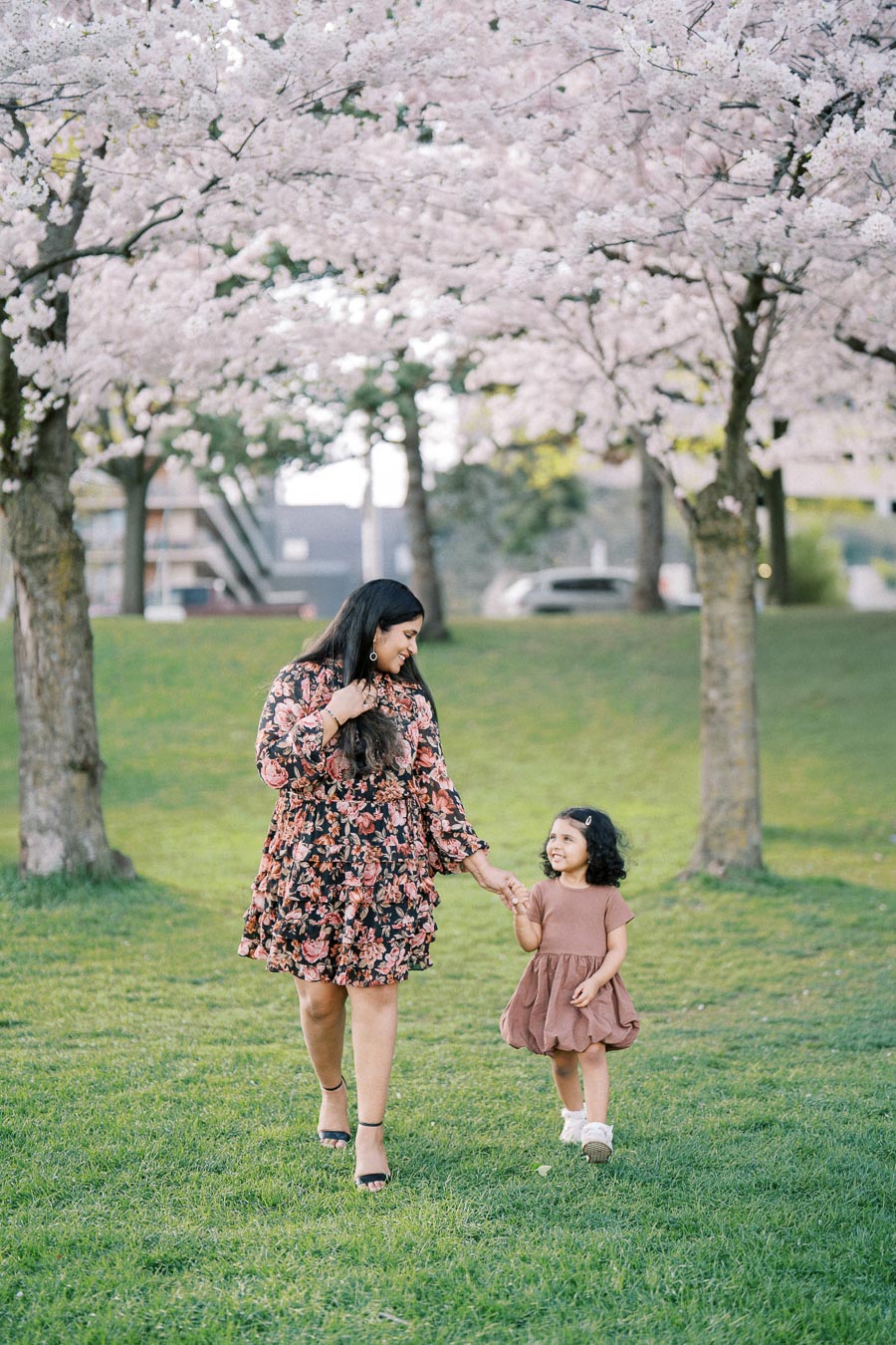 Woman and child walking hand in hand under blooming cherry blossom trees in a park.