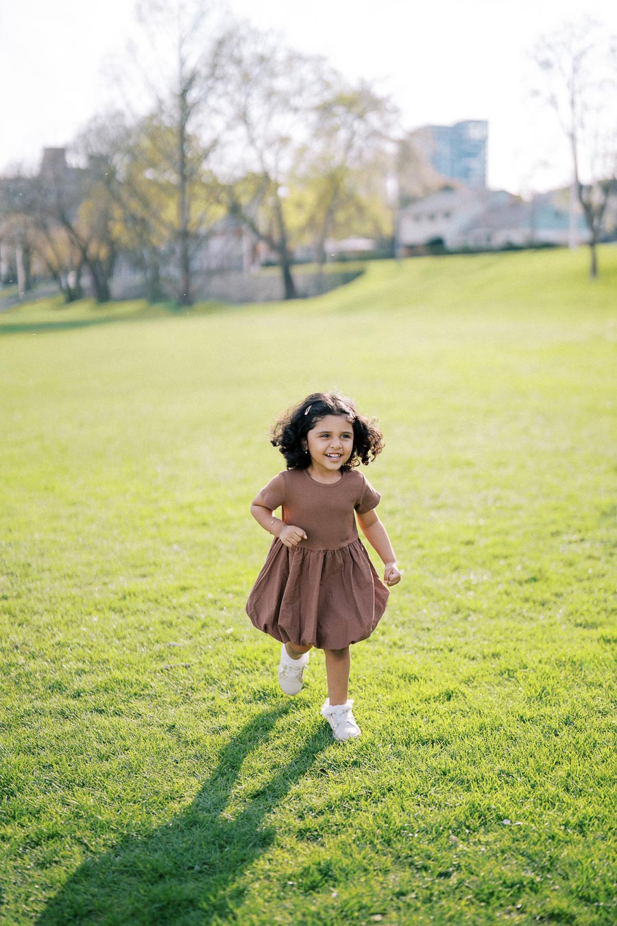 A joyful child in a brown dress running across a sunlit green park, surrounded by trees and buildings in the background.
