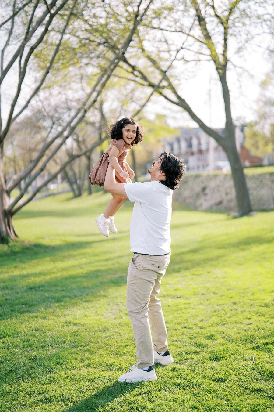 Father lifting his smiling daughter in a park on a sunny day, surrounded by green grass and trees.