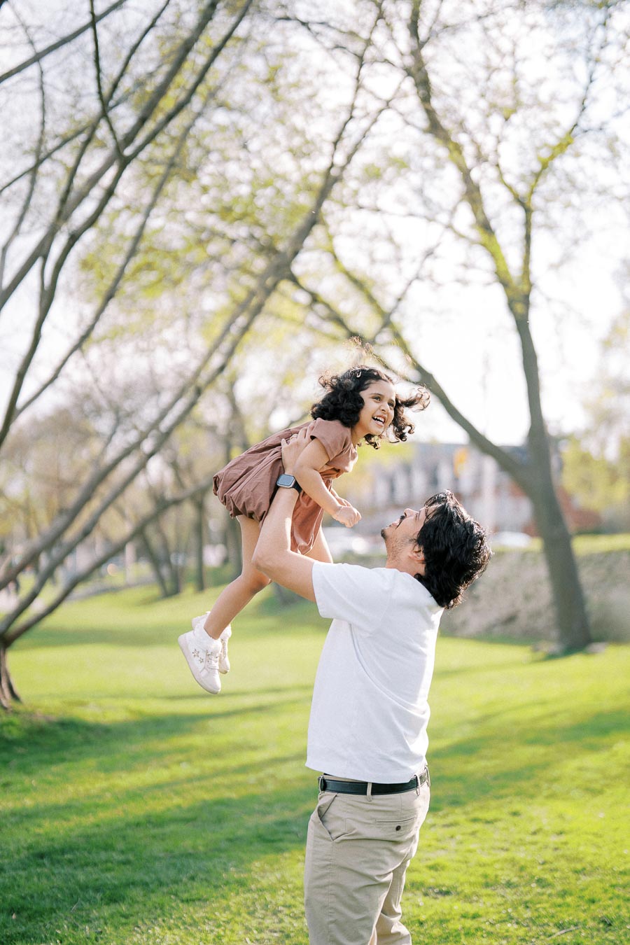 Father lifting daughter joyfully in a sunny park, surrounded by trees and green grass.