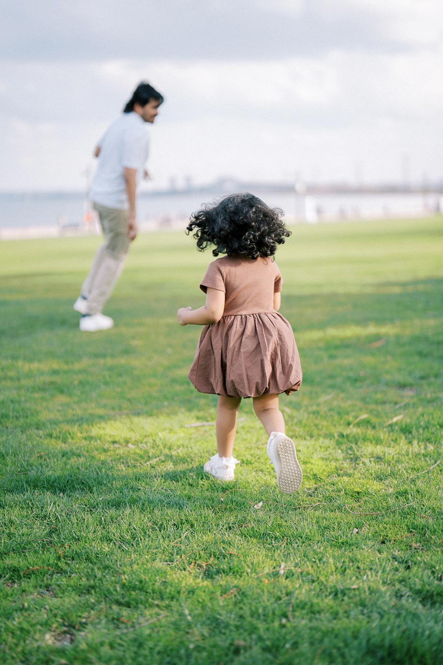 A young child in a brown dress and white shoes running on a green grassy field, with an adult in the background under a cloudy sky, capturing a playful outdoor moment.