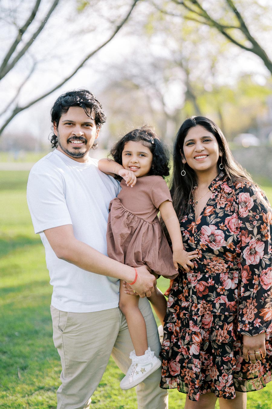 A happy family enjoying a sunny day in the park, with parents smiling and holding their young daughter, surrounded by lush green grass and trees.