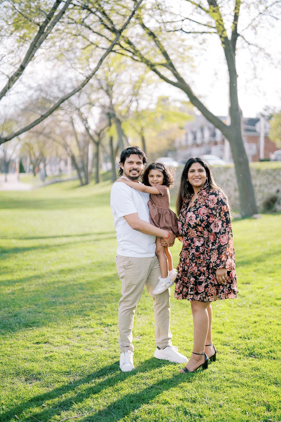 A happy family of three enjoying a sunny day in a park with lush greenery, a father holding his young daughter as the mother stands beside them, showcasing a picturesque and joyful outdoor moment.