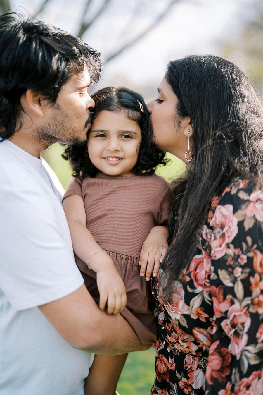 A loving parents kissing their young daughter on the cheeks in a park setting, capturing a joyful family moment.