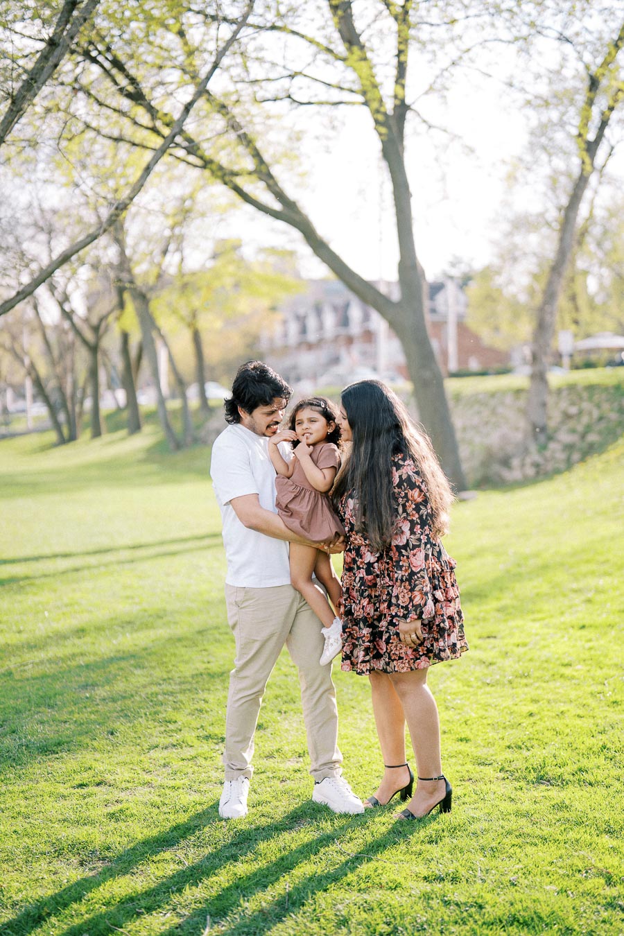 A family enjoying a sunny day at the park, with the parents playfully holding their young daughter while surrounded by lush green trees and grass.