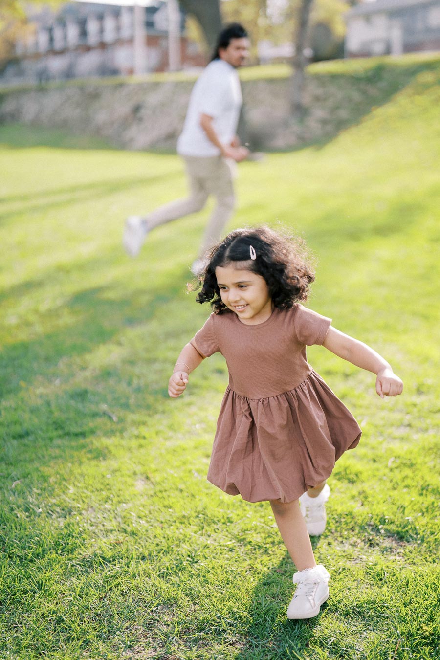 Young girl in a brown dress joyfully running on a grassy field, with a blurred background of an adult following behind, highlighting a playful outdoor scene.