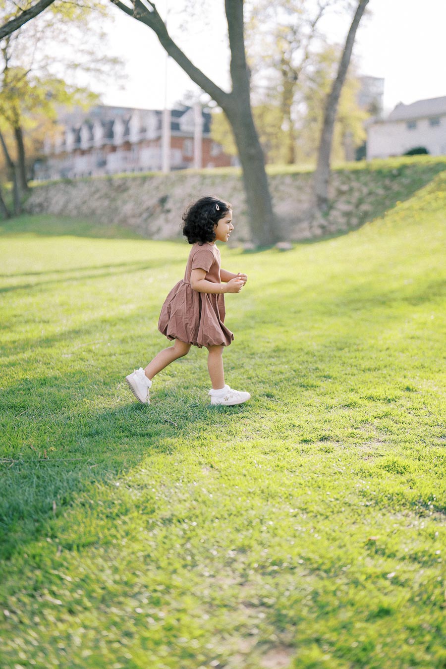 A young child with curly hair, wearing a brown dress and white shoes, happily running on a sunlit grassy field, surrounded by trees and buildings in the background.