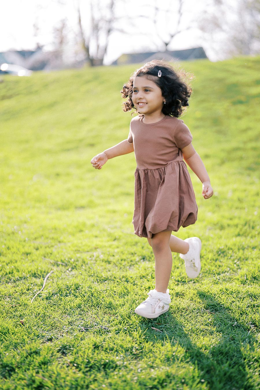 A joyful young girl with curly hair wearing a brown dress and white shoes runs across a lush green field on a sunny day, embodying happiness and playfulness in nature.