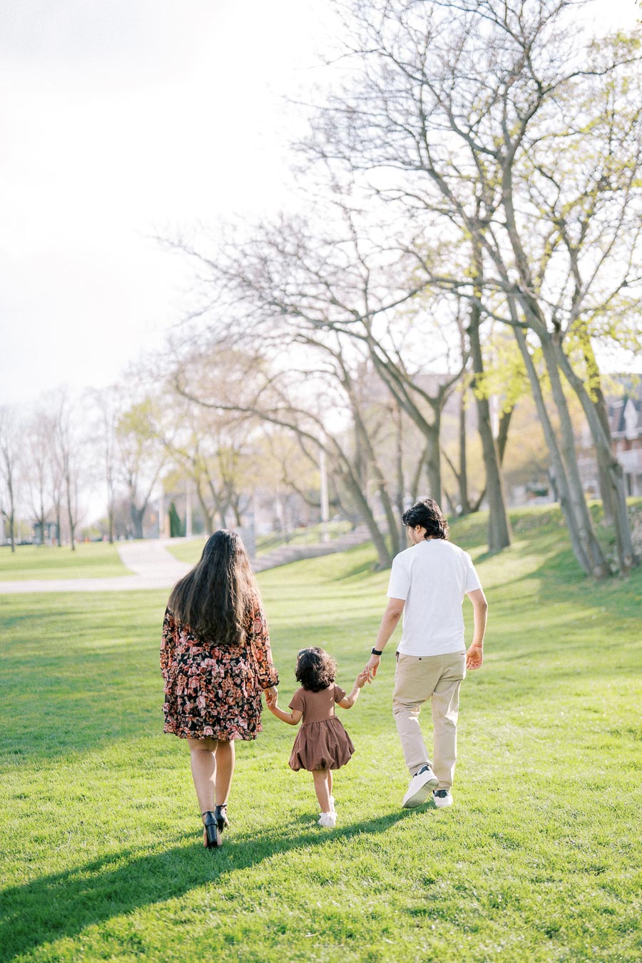 A family walking hand in hand through a sunlit park with trees in the background. The parents are guiding their young child, enjoying a peaceful outdoor moment.