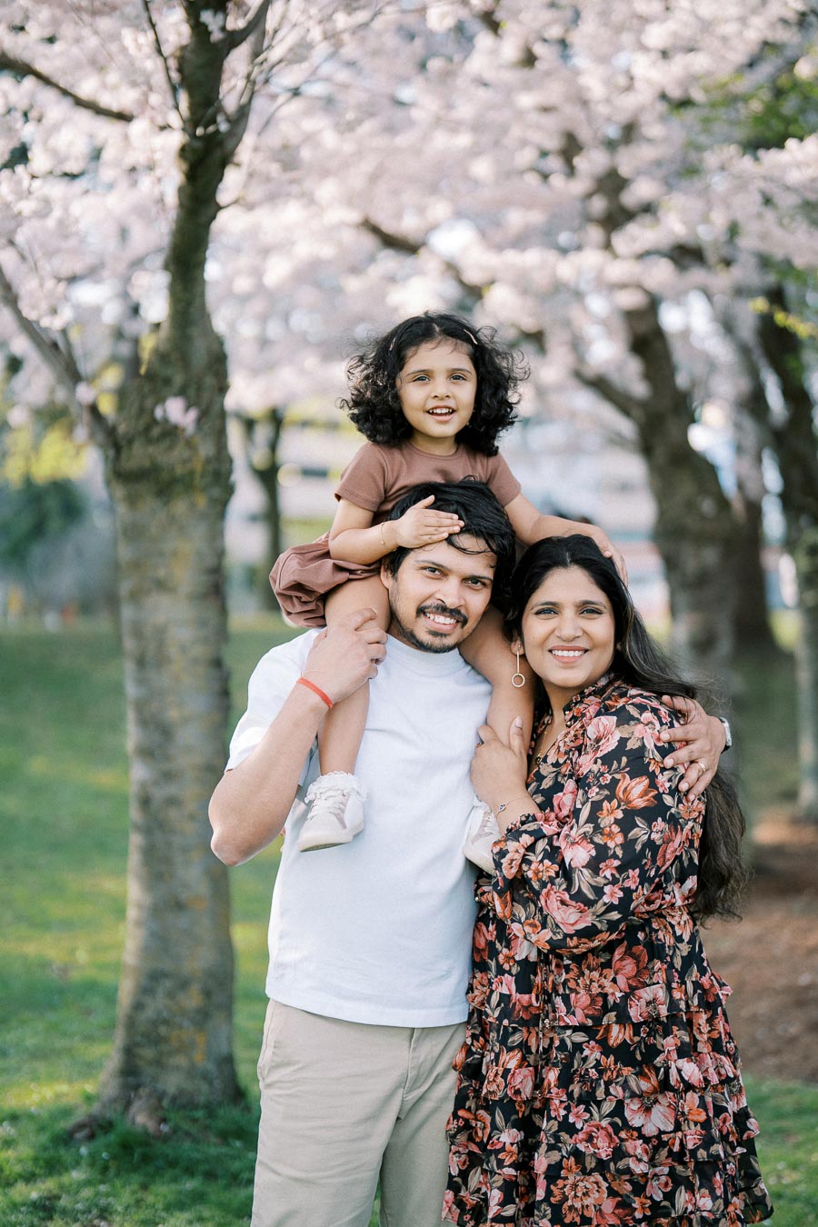 A cheerful family enjoying a day outdoors in a park with cherry blossoms in full bloom, featuring a young girl smiling on her father's shoulders, with both parents smiling and embracing.