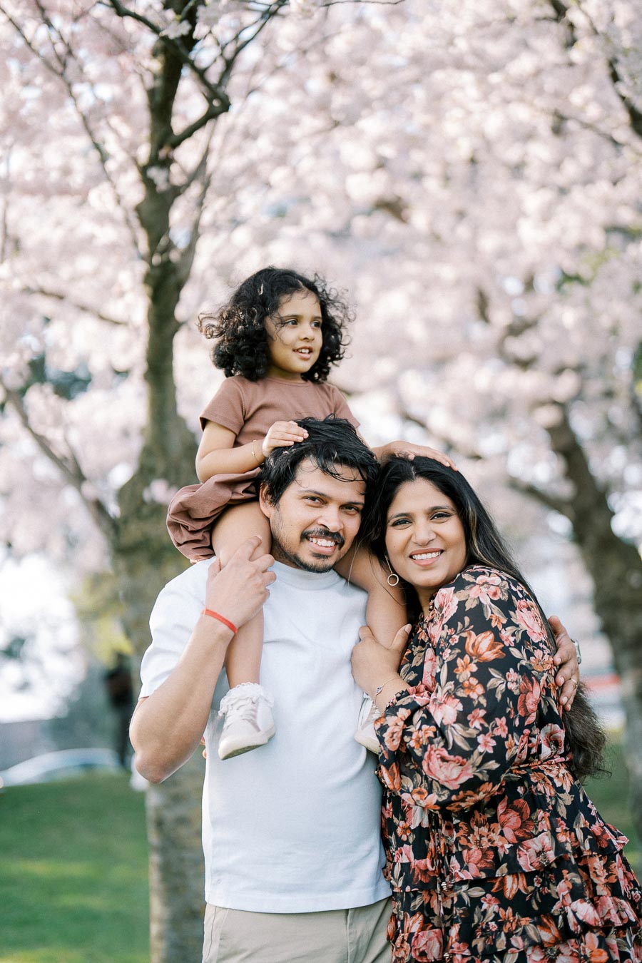 A happy family enjoying a spring day under cherry blossom trees, with a curly-haired child sitting on a man's shoulders and a woman embracing them, all smiling outdoors.