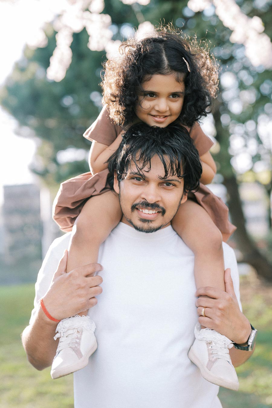 Father smiling while carrying daughter on shoulders in a sunny park.