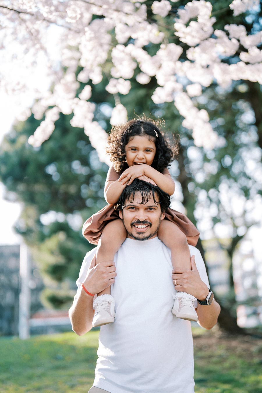 Father with a joyful daughter on his shoulders under cherry blossom trees in a park.