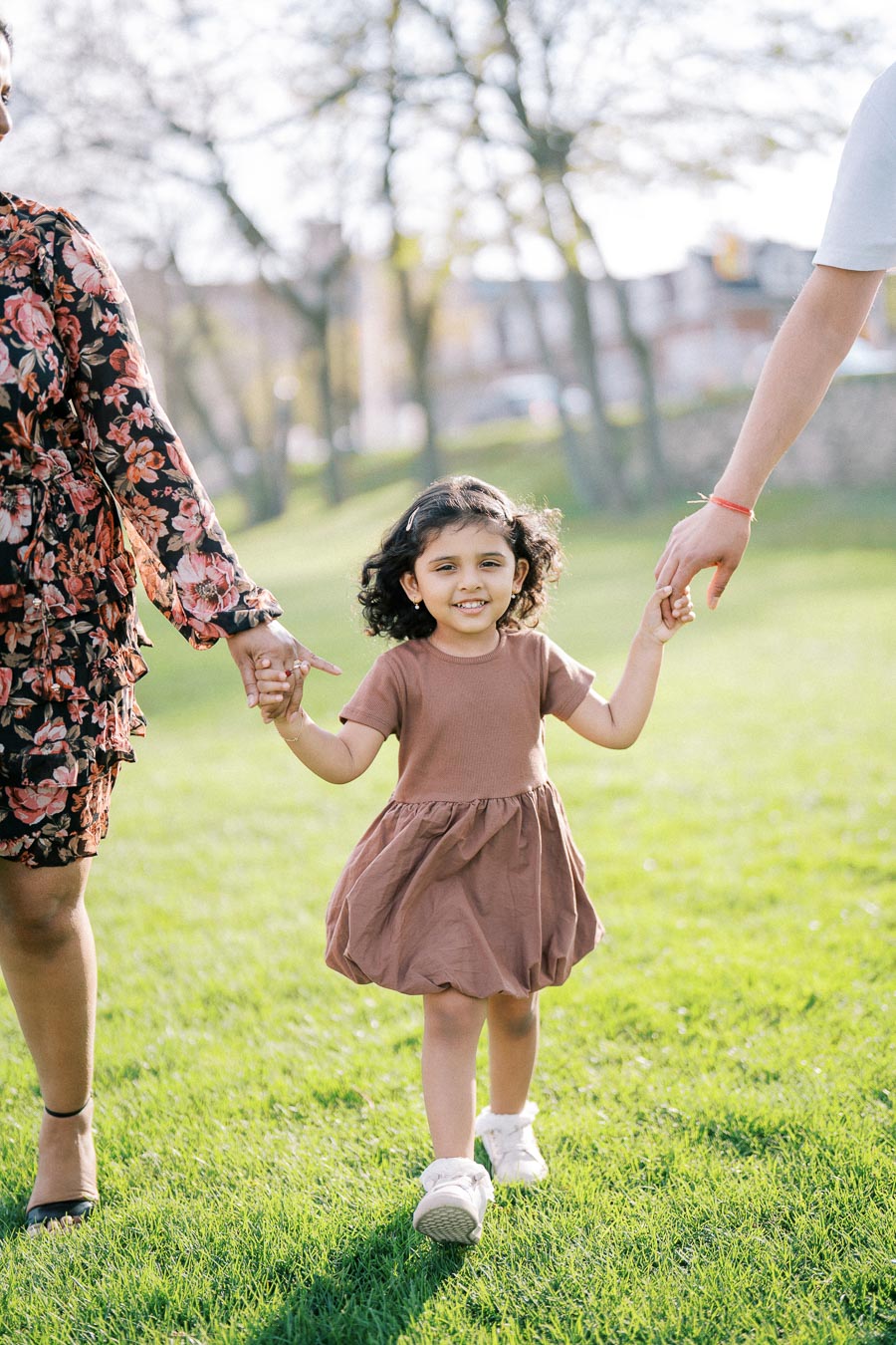 A young girl in a brown dress smiles as she walks hand in hand with two adults in a sunlit park, surrounded by green grass and trees.