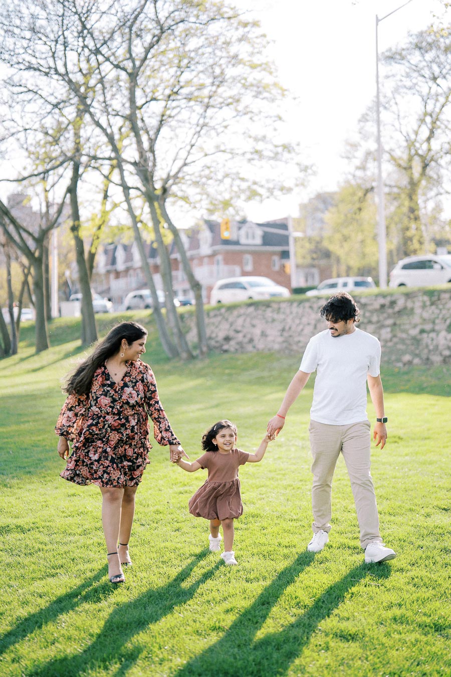 A happy family walks hand-in-hand across a sunny park, with fresh greenery and trees in the background, capturing a joyful moment outdoors.