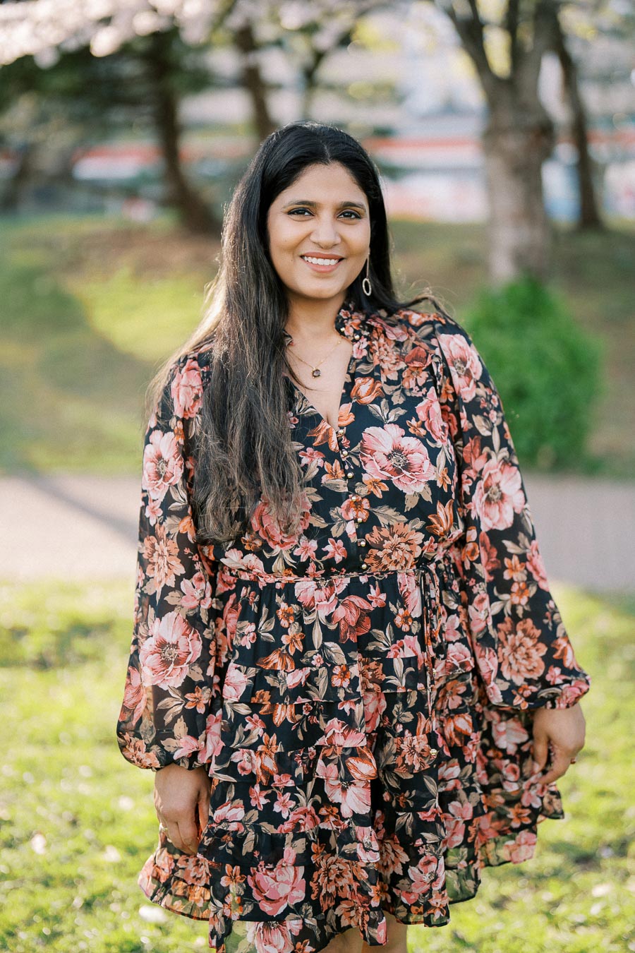A woman in a floral dress standing outdoors in a park, smiling, with green grass and trees in the background.