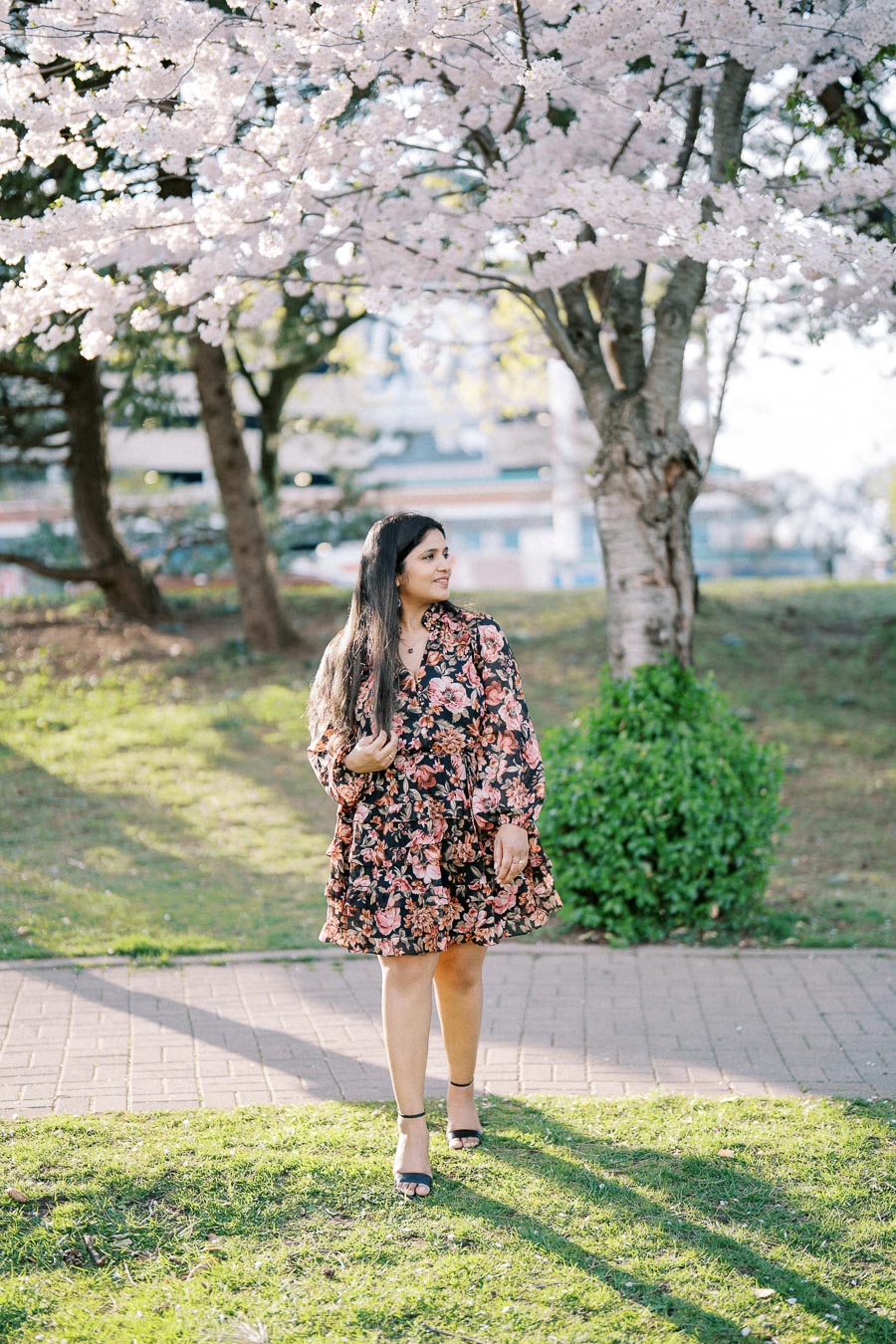 A woman in a floral dress stands under a cherry blossom tree in a sunlit park, surrounded by greenery and a brick path in the background.