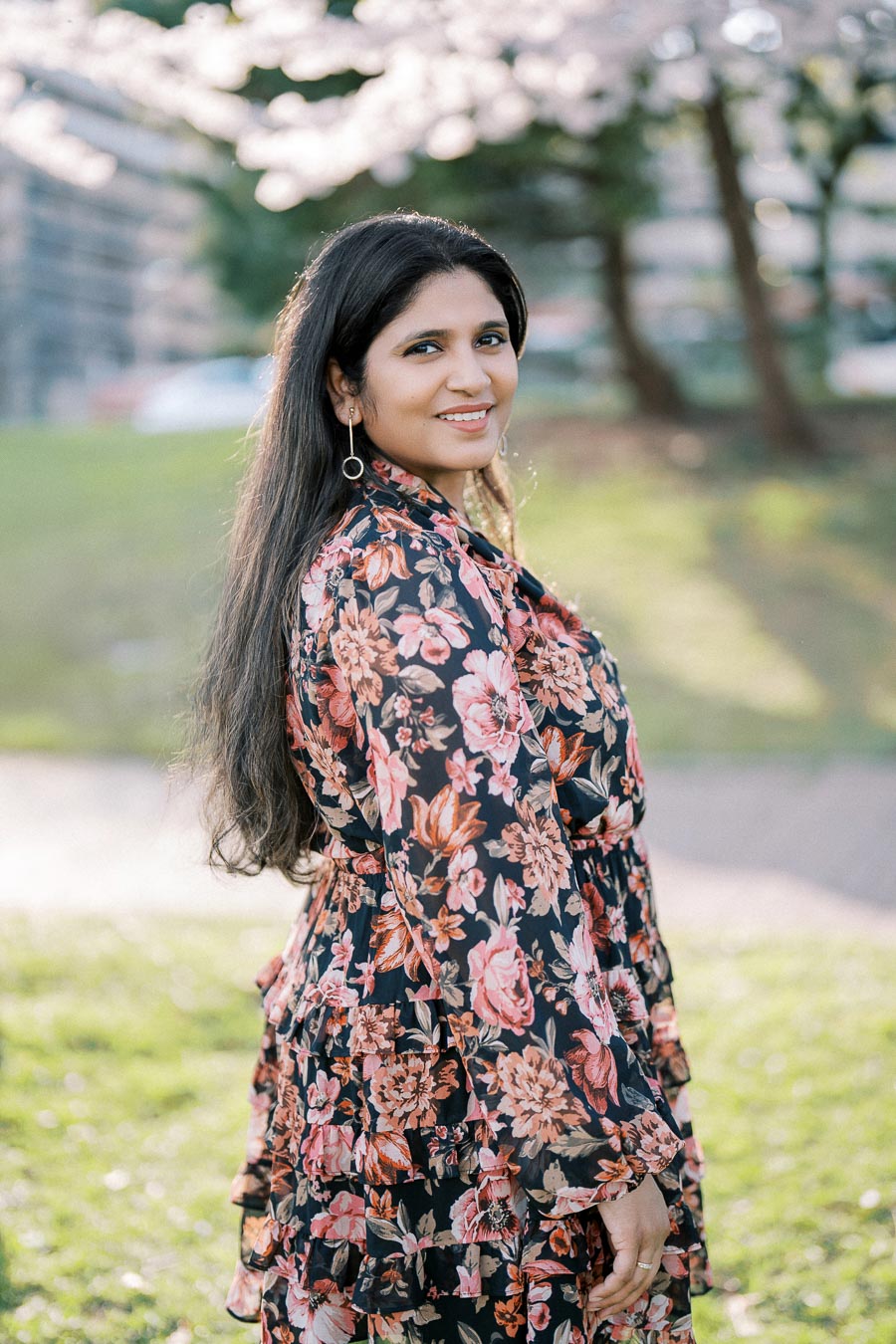 A woman in a floral dress smiles while standing in a park under cherry blossom trees, with soft sunlight illuminating the scene.