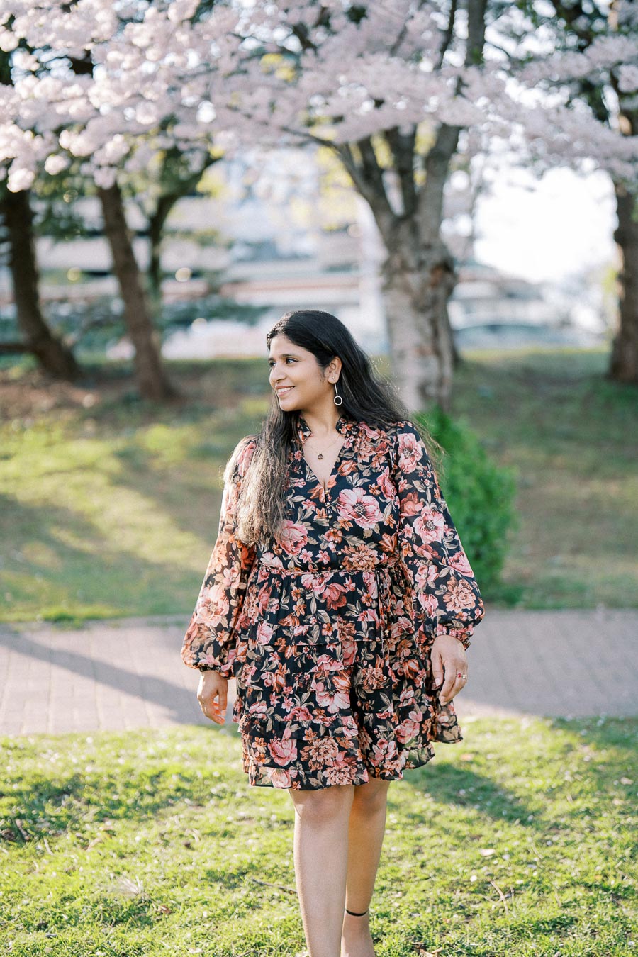 A woman in a floral dress walks through a park with blooming cherry blossom trees in the background, enjoying a sunny day.