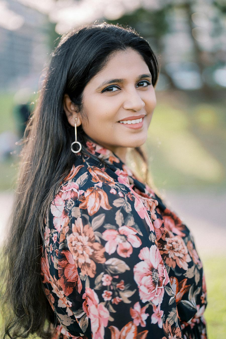 Smiling woman with long dark hair wearing a floral-patterned blouse and hoop earrings, standing outdoors with a blurred green background.