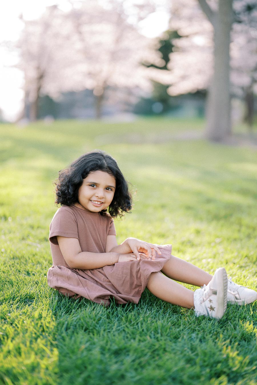 A young child sitting on green grass, smiling, wearing a brown dress and white sneakers. Cherry blossoms softly blurred in the background, capturing a serene spring day outdoors.