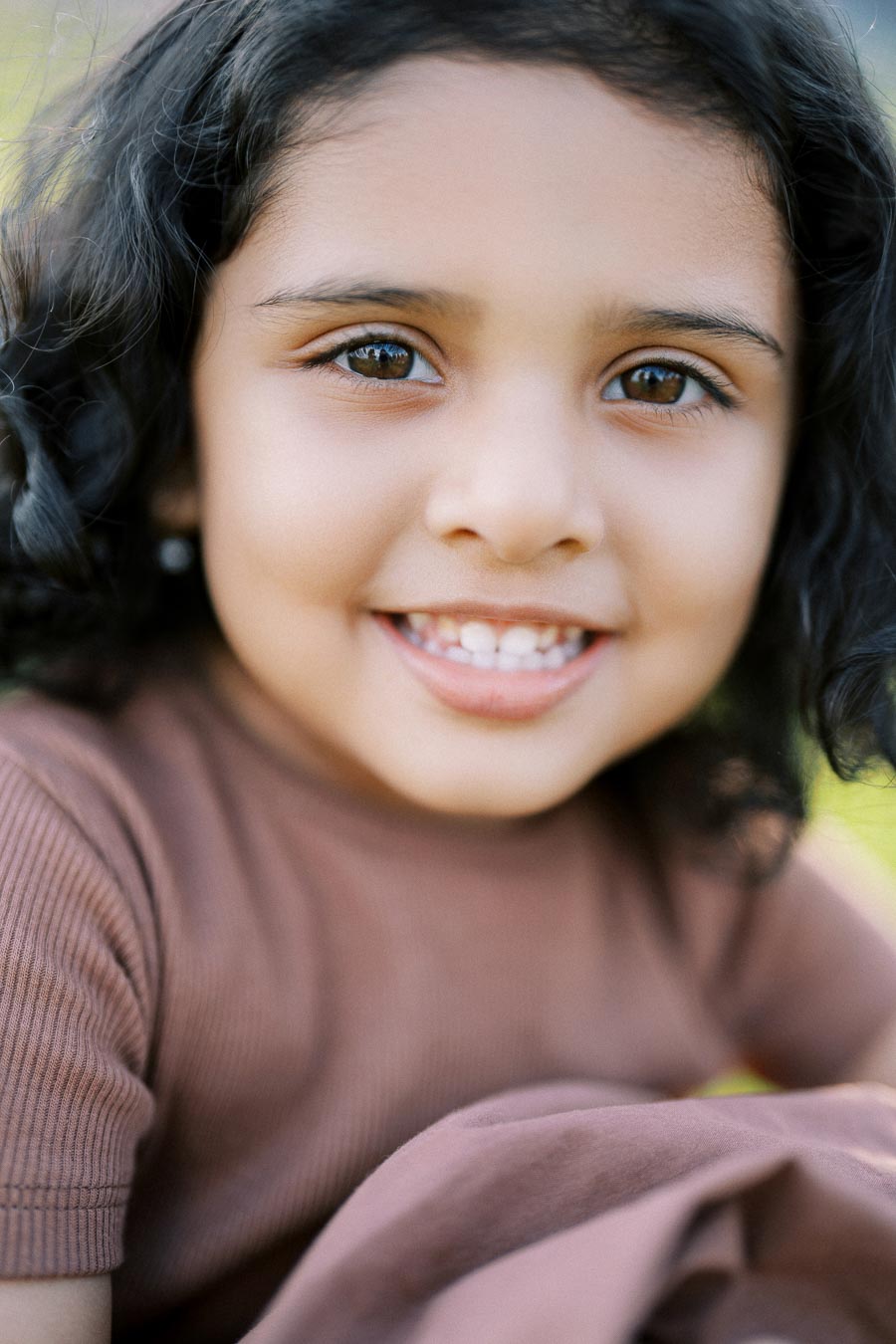 Close-up of a smiling child with curly hair, wearing a brown shirt, against a blurred outdoor background
