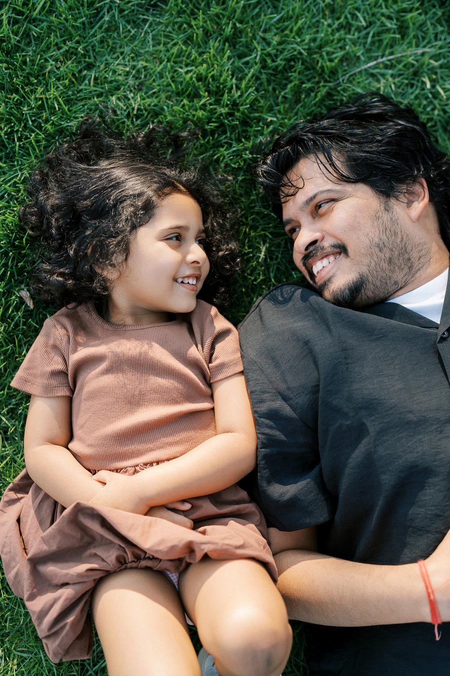 Father and daughter lying on grass, smiling and looking at each other, enjoying a sunny day outdoors.