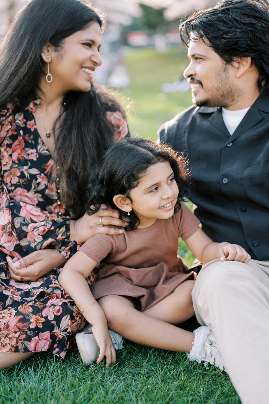 Happy family enjoying a day outdoors, mother and father sitting on the grass with their young daughter in the middle, smiling and wearing casual clothes.